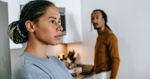 Woman in the foreground looking defensive while her partner in the background speaks to her in the kitchen