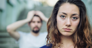 Woman looking angrily at the camera while a man in the background puts his hands on top of his head